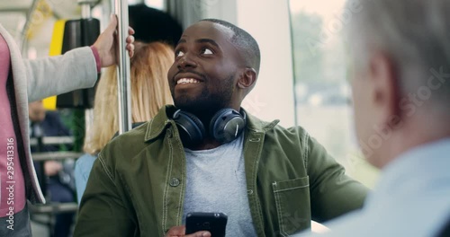African American handsome and cheerful polite young man holding smartphone and sitting in the tram while going somewhere, then standing up and giving his sit to the old Caucasian woman.