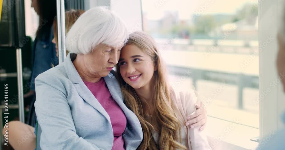 Portrait shot of the pretty and happy Caucasian grandmother and her cute granddaughter sitting in the tram and hugging while going somewhere with grandfather who siting in front of them. Rear. Portrait shot of the pretty and happy Caucasian grandmother and her cute granddaughter sitting in the tram and hugging while going somewhere with grandfather who siting in front of them. Rear.