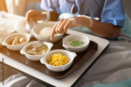 Woman wearing a blue patient hospital gown, eating food for patients in on the patient bed in the hospital