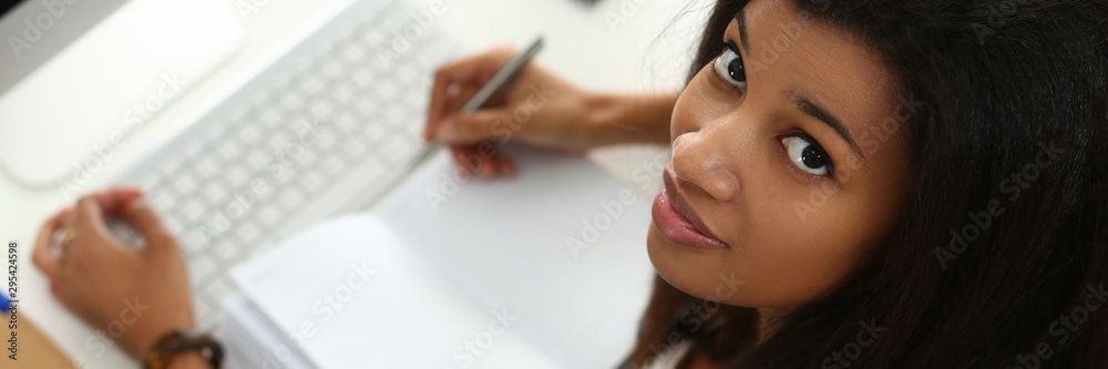 Young attractive smiling black teenager girl sit at office workplace look in camera portrait. Freelance creative person desktop pc blank synopsis notepad page exam preparation concept