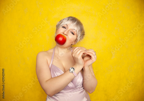 charming funny plus size woman with short hair shows the heart symbol with her hands and holds a red Apple in her mouth.