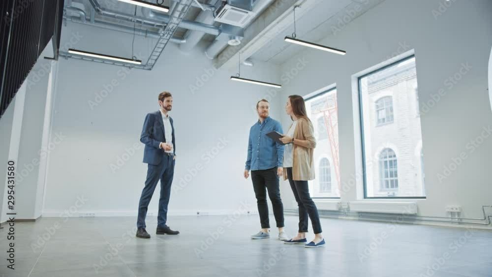 Real Estate Agent Showing a New Empty Office Space to Young Male and Female Hipsters. Entrepreneurs Meet the Broker with a Tablet and Discuss the Facility They Wish to Purchase or Rent. Real Estate Agent Showing a New Empty Office Space to Young Male and Female Hipsters. Entrepreneurs Meet the Broker with a Tablet and Discuss the Facility They Wish to Purchase or Rent.