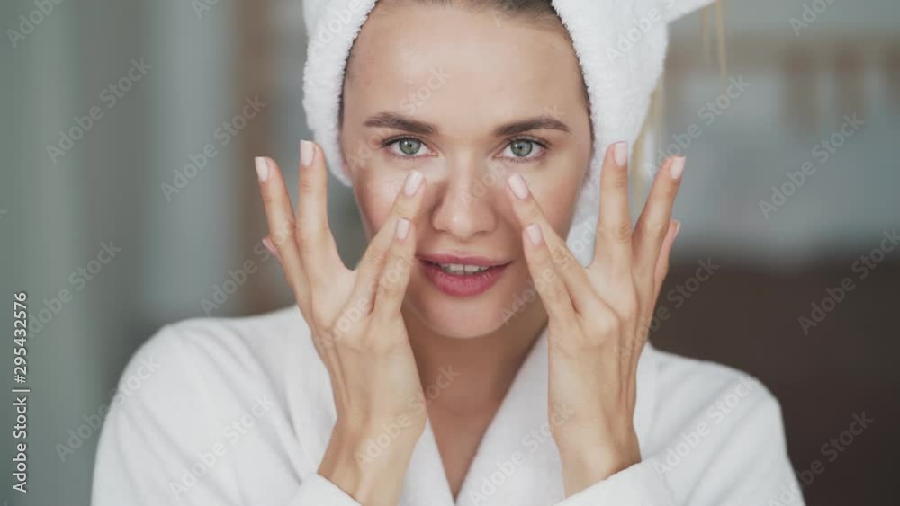 Portrait of woman in bathrobe, with towel on head applies cream on her face Portrait of woman in bathrobe, with towel on head applies cream on her face