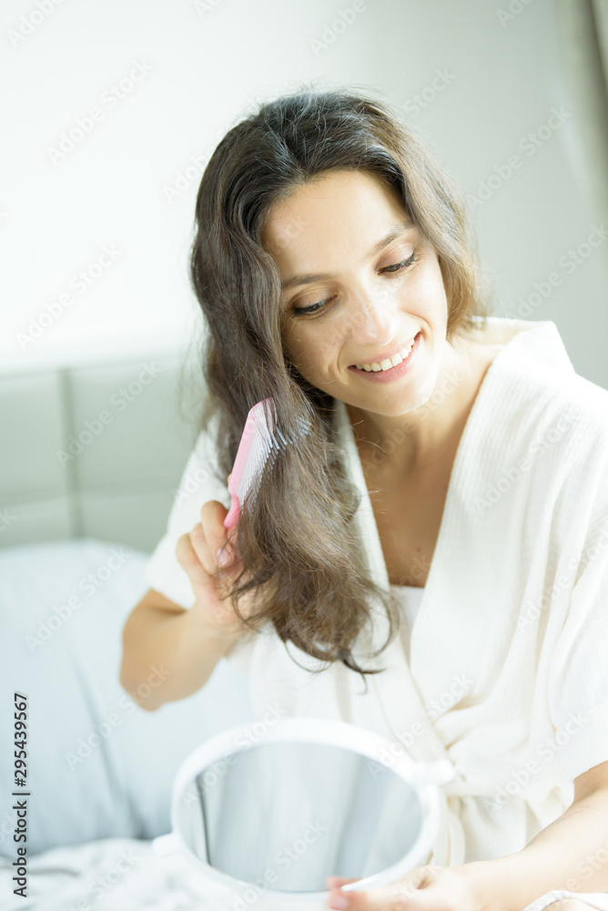 A beautiful woman wearing a towel and a white bathrobe has to look a mirror for combing hair with a pink comb and on the bed at a condominium in the morning. A beautiful woman wearing a towel and a white bathrobe has to look a mirror for combing hair with a pink comb and on the bed at a condominium in the morning.