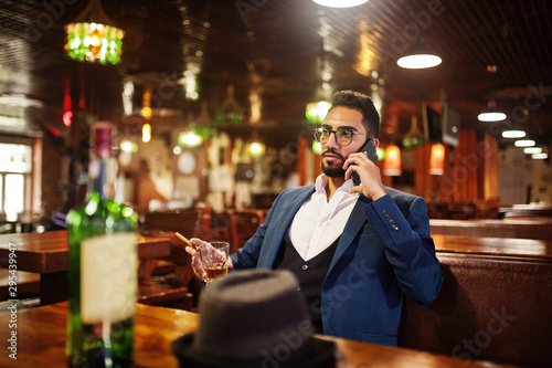 Handsome well-dressed arabian man with glass of whiskey and cigar hold mobile phone, posed at pub.