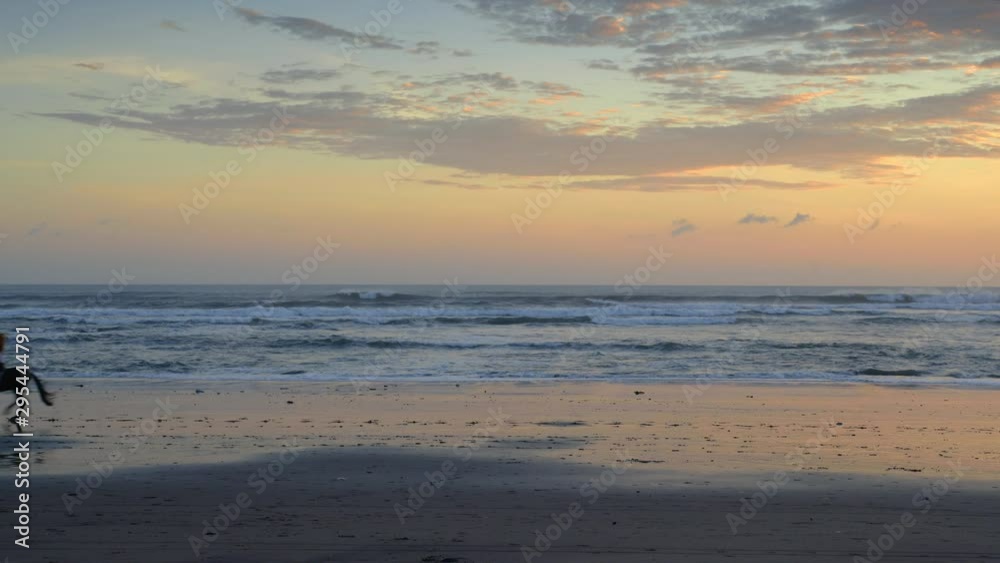 A young woman is riding and galloping a big black horse on a beach during colorful sunset and high waves A young woman is riding and galloping a big black horse on a beach during colorful sunset and high waves