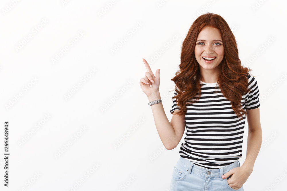 Carefree smiling cute redhead woman in striped t-shirt smiling delighted, discuss cool new offer, pointing upper left corner, indicate copy space promo, grin satisfied, standing white background Carefree smiling cute redhead woman in striped t-shirt smiling delighted, discuss cool new offer, pointing upper left corner, indicate copy space promo, grin satisfied, standing white background