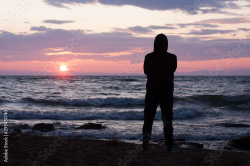 Solitude, loneliness, mindfulness concept. Silhouette of young man standing on edge of beach and looking to horizon of sea waves. Dreaming and thinking alone person at sunset seashore. 