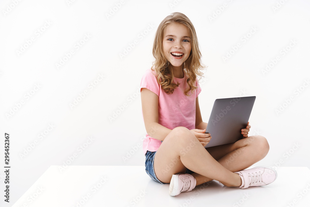 Fototapeta premium Pretty cheerful little kid girl sit crossed legs, hold laptop, studying at home, excited learn new information, studying via internet, contact language teacher video-call lesson, white background