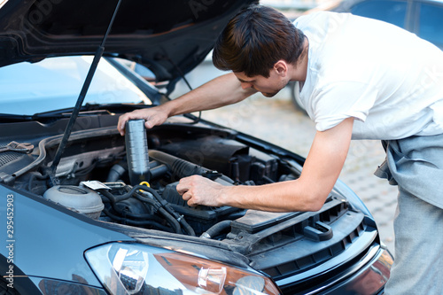 Young brunette man is changing oil in auto with open hood. Driver is repairing automobile on street road. Vehicle breakdown on way concept. Repairman mechanic is servicing car in workshop.