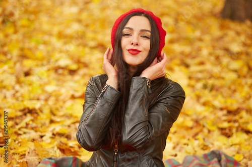 Image of attractive female, brunette woman dresses stylish beret and leather jacket, looking at camera and smiling, posing outdors, being in good mood, has pleasant appearance, keeps hands near neck.