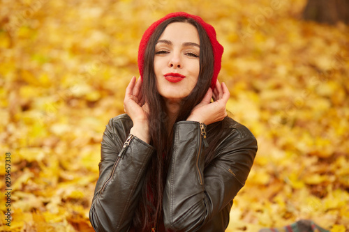 Horizontal shot of beautiful woman showing kiss gesture and smiling to camera, posing outdoor on autumn park background, charming girl dresses red beret and leather jacket, surrounded yellow leaves.