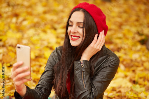 Fashion portrait of young woman outdoor in autumn park or foreest, lady wearing lather jacket, and red beret, taking self picture viaher modern telephone, charming girl surrounded with yellow leaves. photo