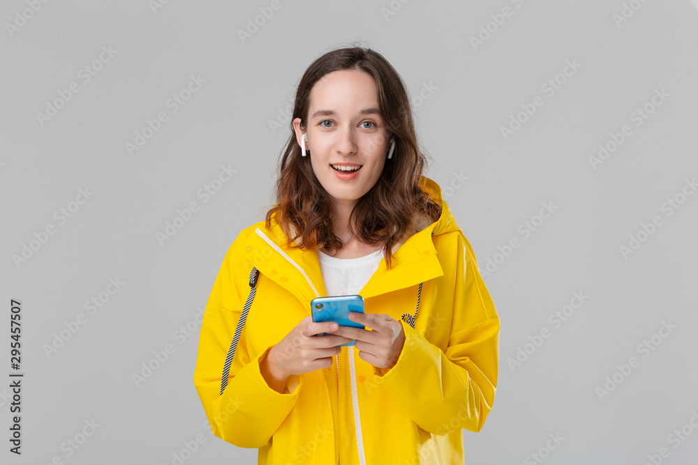 Smiling brunette woman in a yellow raincoat is reading notification on the smartphone. Model wears wireless earbuds. Concept of communication. Smiling brunette woman in a yellow raincoat is reading notification on the smartphone. Model wears wireless earbuds. Concept of communication.
