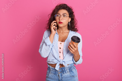Half length shot of astonished Caucasian woman, posing with widely opened mouth, hears shocking news from her friend, usesing her mobile phone, dressed casually, posing indoor against pink wall.