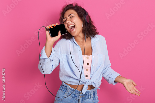 Picure of woman listening to music on headphones, dancing and singigng, using her smart phone like microphone. Closeup portrait of beautiful girl wearing blue shirt and jeans, lady having fun.