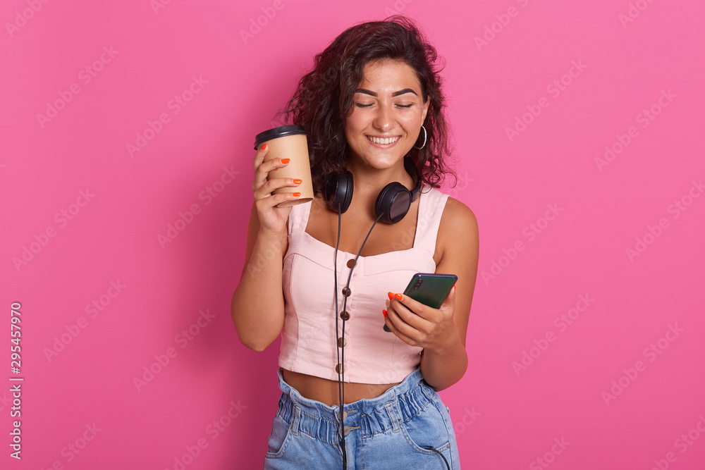 Cheerful girl having coffee break, reading notification on her mobile phone, updates favourite app, typing message toher friend and smiles, wearing rosetop and jeans, isolated over pink background. Cheerful girl having coffee break, reading notification on her mobile phone, updates favourite app, typing message toher friend and smiles, wearing rosetop and jeans, isolated over pink background.