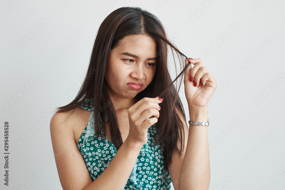 Beautiful Asian brunette girl with sad expression on face looks at her dull, dry, brittle dyed hair unhappy with their quality isolated on white background with copy space Beautiful Asian brunette girl with sad expression on face looks at her dull, dry, brittle dyed hair unhappy with their quality isolated on white background with copy space