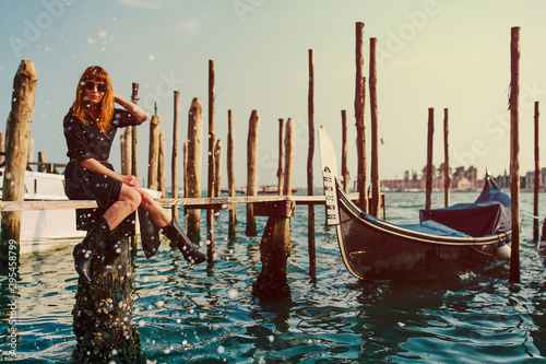 Redhead girl with long black dress, sunglasses and cowboy boots sitting on Gondola boards in Venice
