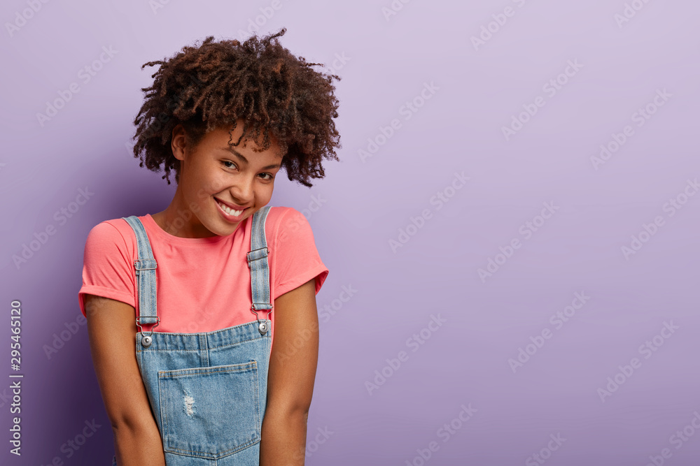 Delighted African American woman has cheerful shy face expression,  expresses positive emotions, enjoys compliment, wears pink t shirt and denim  sarafan, models over purple wall, copy space aside Stock Photo | Adobe