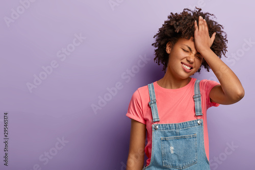 Disappointed troublesome woman keeps palm on forehead, regrets did something wrong, face problematic situation, dressed in fashionable otfit, poses over purple wall, forgets about important task