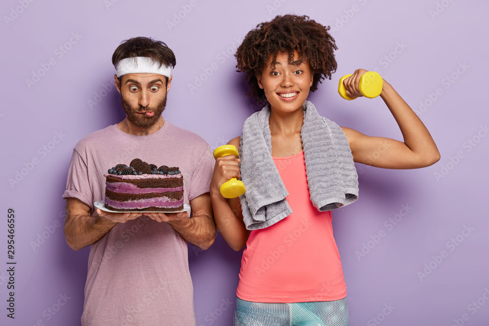 Surprised young Afro man stares at delicious cake, wears white headband, feels temptation, happy woman works on biceps, raises weights, leads sporty lifestyle, stand against purple background.