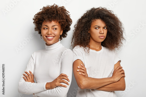 Smiling dark skinned woman keeps hands crossed, stand shoulder to shoulder with best friend who has gloomy upset expression, wear white clothes, express different emotions. Ethnicity and relations