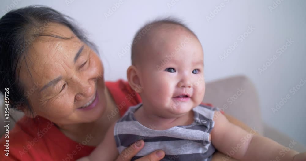 Close up slow motion of happy Chinese grandmother holding playing with her baby infant granddaughter at home old and young together Close up slow motion of happy Chinese grandmother holding playing with her baby infant granddaughter at home old and young together