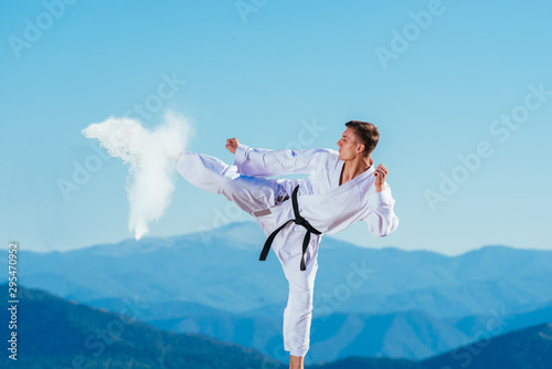 Fit karate athlete kicking a cup filled whit fluor causing a big splash while wearing a white kimono on top of a mountain on a sunny day.
