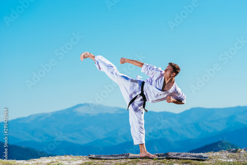 Karate man in a kimono performs a side leg-foot kick(Mae-geri) while standing on the green grass on top of a mountain.