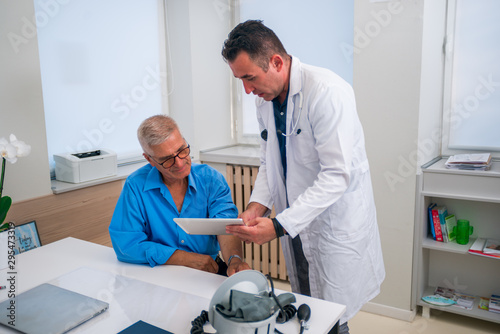 Middle aged caucasian doctor explains to his senior patient his blood pressure readings while standing next to him.