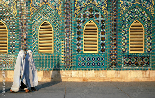 The Blue Mosque in Mazar-i-Sharif, Balkh Province in Afghanistan. Two women wearing white burqas (burkas) walk past a wall of the mosque adorned with colorful tiles and mosaics. Northern Afghanistan. photo