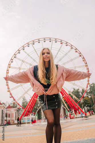 young casual blonde girl with long hair is standing near big red Ferris wheel and pretends to hold on to him, lifestyle concept, free space from above