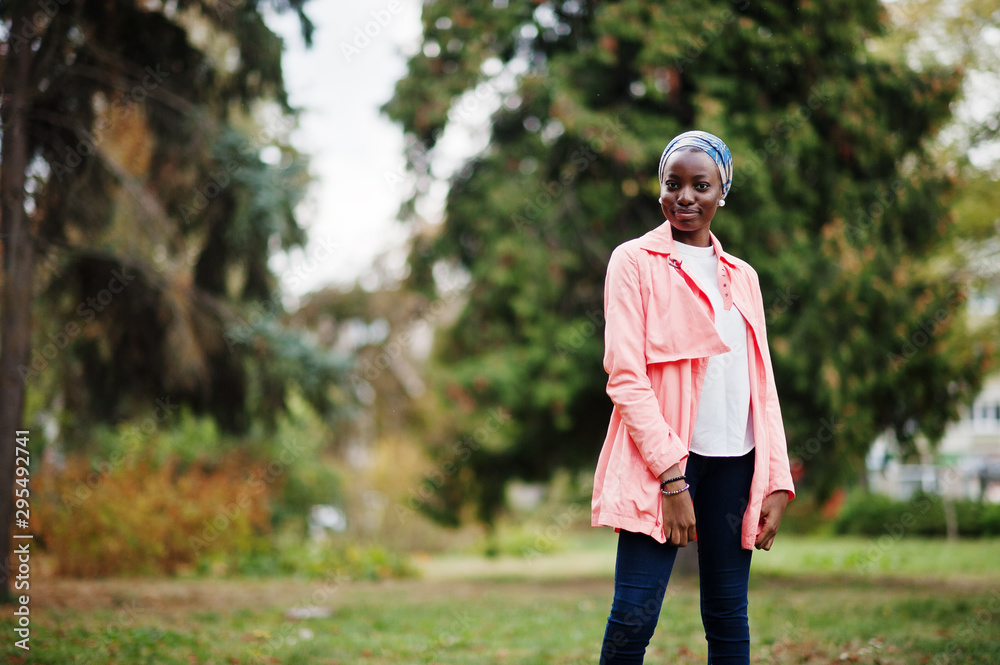 Obraz premium Young modern fashionable, attractive, tall and slim african muslim woman in hijab or turban head scarf and pink coat posed at park.