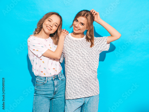 Two young beautiful smiling blond hipster girls in trendy summer colorful T-shirt clothes. Sexy carefree women posing near blue wall. Positive models having fun