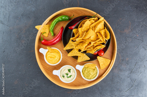 Nachos - yellow corn chips with various sauces in bowls: guacamole, cheese sauce, white sauce, on a dark background. Mexican food concept. The view from the top.