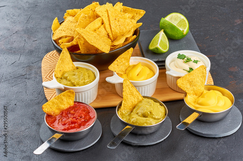 Nachos - yellow corn chips with various sauces in bowls: guacamole, cheese sauce, salsa sauce, white sauce, on dark background. Mexican food concept.