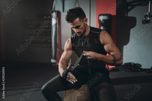 Portrait concentrated brutal athletic guy wraps hands using the professional bandages while sitting on the chair in the gym class