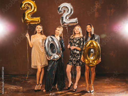 Beautiful Women Celebrating New Year. Happy Gorgeous Girls In Stylish Sexy Party Dresses Holding Gold and Silver 2020 Balloons, Having Fun At New Year's Eve Party. Holiday Celebration.Charming Models