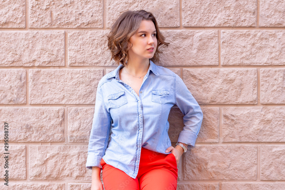 female portrait. beautiful young girl stands on a background of a beige brick wall with stucco. red pants and a denim shirt. beige background female portrait. beautiful young girl stands on a background of a beige brick wall with stucco. red pants and a denim shirt. beige background