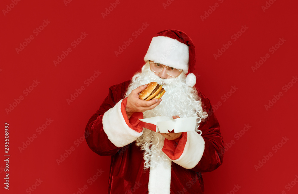 Funny man in Santa Claus costume stands on red background with burger in hand, looks at fast food with surprised face. Shocked Santa isolated on red background with sandwich in hand. Funny man in Santa Claus costume stands on red background with burger in hand, looks at fast food with surprised face. Shocked Santa isolated on red background with sandwich in hand.
