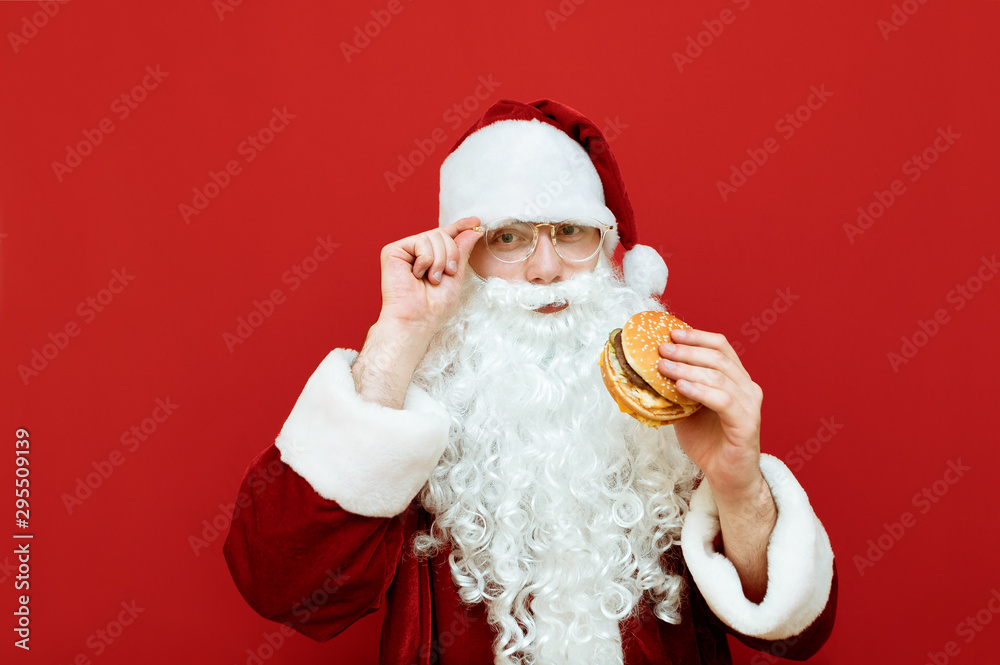 Positive Santa Claus with white beard and glasses stands on a red background, looks in camera and smiles, holds a burger in his hand. Portrait of Santa isolated on red background with burger in hand