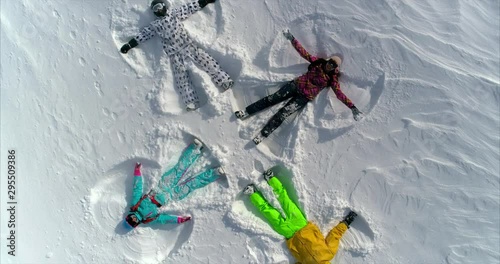 AERIAL CLOSE UP DISTANCING: very bright and cheerful snow angels lie on a snow field four, waving their arms and legs and throw snow up. the camera flies up and spins. winter concept. SLOW MOTION
