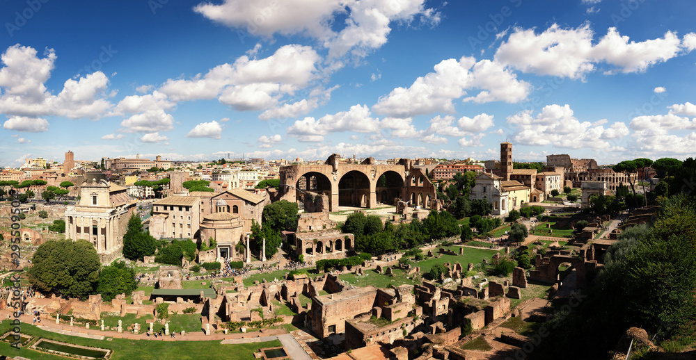 Panoramic photo of the ruins of the Roman forums. On the background the roofs of Roman houses and the blue sky with clouds; on the right there is the Colosseum.