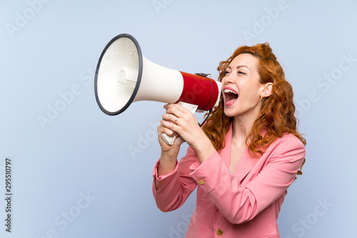 Redhead woman in suit over isolated blue wall shouting through a megaphone