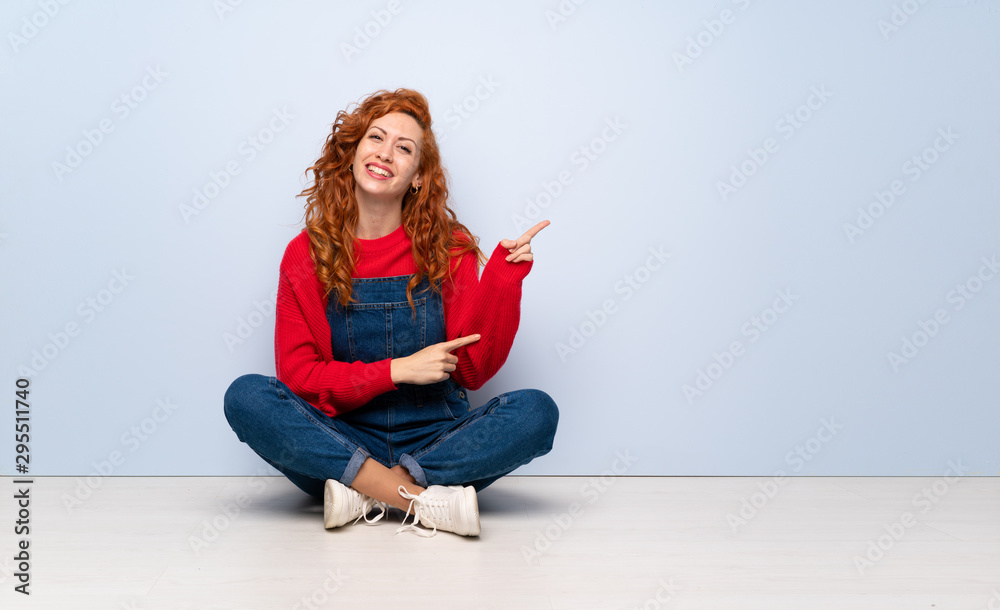 Redhead woman with overalls sitting on the floor pointing finger to the side Redhead woman with overalls sitting on the floor pointing finger to the side