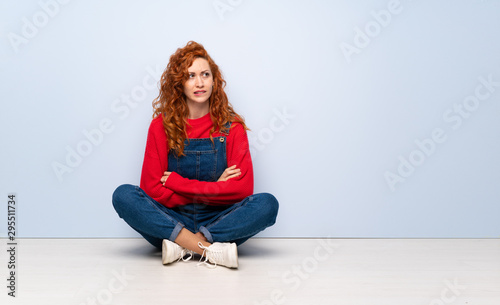 Redhead woman with overalls sitting on the floor with confuse face expression