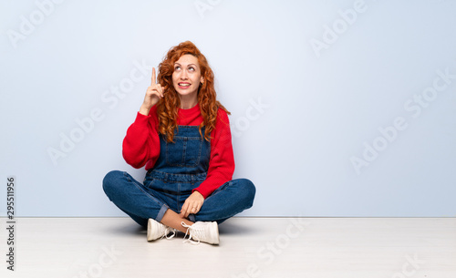 Redhead woman with overalls sitting on the floor intending to realizes the solution while lifting a finger up © luismolinero