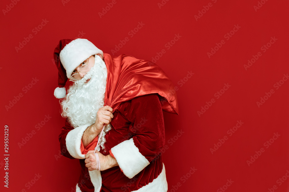 Portrait of Santa Claus with heavy bag with gifts on his back, isolated on red background, looking into camera with serious face. Santa carries bag on red background. Christmas concept. Copyspace Portrait of Santa Claus with heavy bag with gifts on his back, isolated on red background, looking into camera with serious face. Santa carries bag on red background. Christmas concept. Copyspace