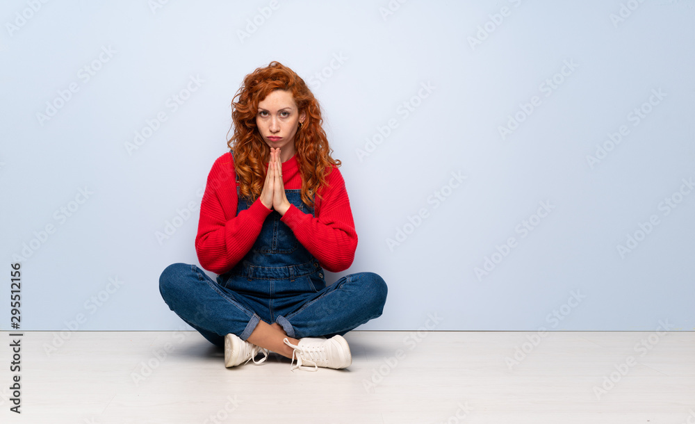 Redhead woman with overalls sitting on the floor keeps palm together. Person asks for something Redhead woman with overalls sitting on the floor keeps palm together. Person asks for something